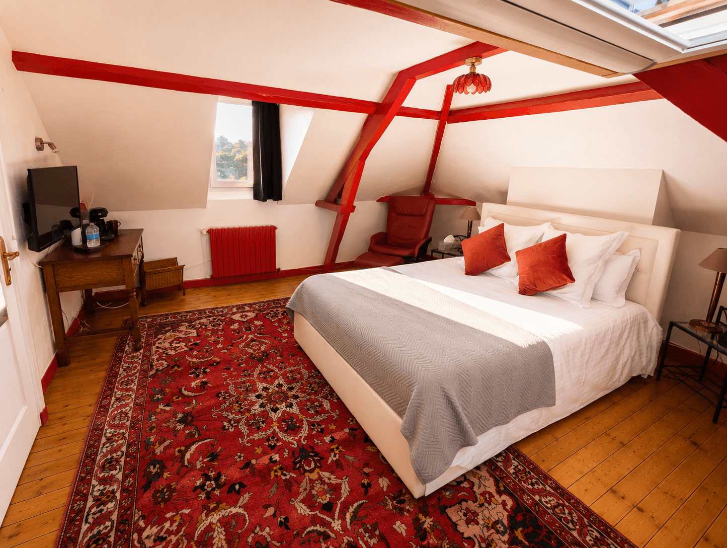 Chambre mansardée aux poutres rouges, lit blanc et tapis persan